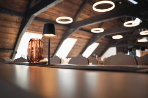 timmungsvolle Detailaufnahme eines Tisches mit einer kleinen, modernen schwarzen Tischlampe und einem bernsteinfarbenen Deko-Gefäß aus Glas. Im Hintergrund sind unscharf die Dachschrägen zu erkennen.